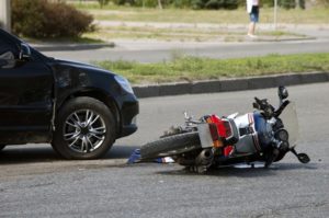 A motorbike on its side on the road in front of a black car.