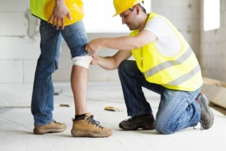A construction worker applies a bandage to a colleague with a knee injury