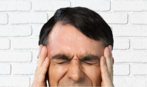 A man in front of a white brick wall holding his temples on his face with his hands and scrunching his face with his eyes closed.