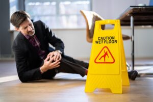 A man lying on the floor holding his knee in pain after slipping next to a wet floor sign.