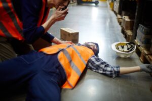 A man in an orange workers vest lays unconscious on the floor of a warehouse