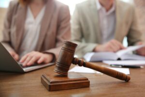 Two people, one in a light brown suit and one in a cream suit, sitting behind a desk with a judge's gavel sat on it.