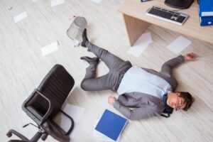An office worker unconscious on the floor with their office chair tipped over.