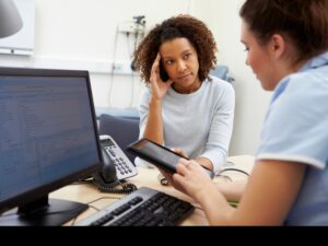 A GP shows a tablet to a patient, who has one hand on their head.