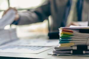 A pile of different colour coordinated documents stacked on top of each other on a desk. Blurred in the background is business person in a suit sat at the desk.