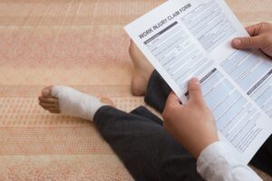 A man with bare feet and one bandaged foot sat on the floor, he is holding a work injury claim form. 