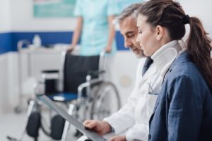 A doctor examining the medical records of his badly injured patient. A nurse stands ready with their wheelchair in the background.
