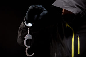A hooded and masked person with handcuffs dangling from one hand. 