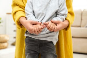 A woman in a yellow coat stands behind a child holding their hands.