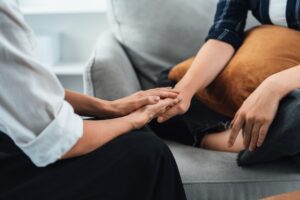 A woman sits cross-legged with a pillow on her lap as another woman holds her hand.