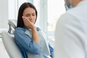 Woman sits in an examination chair holding her face in pain with a dentist nearby.
