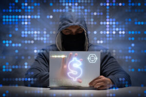 A masked and hooded man at a transparent laptop with a dollar sign on it. 